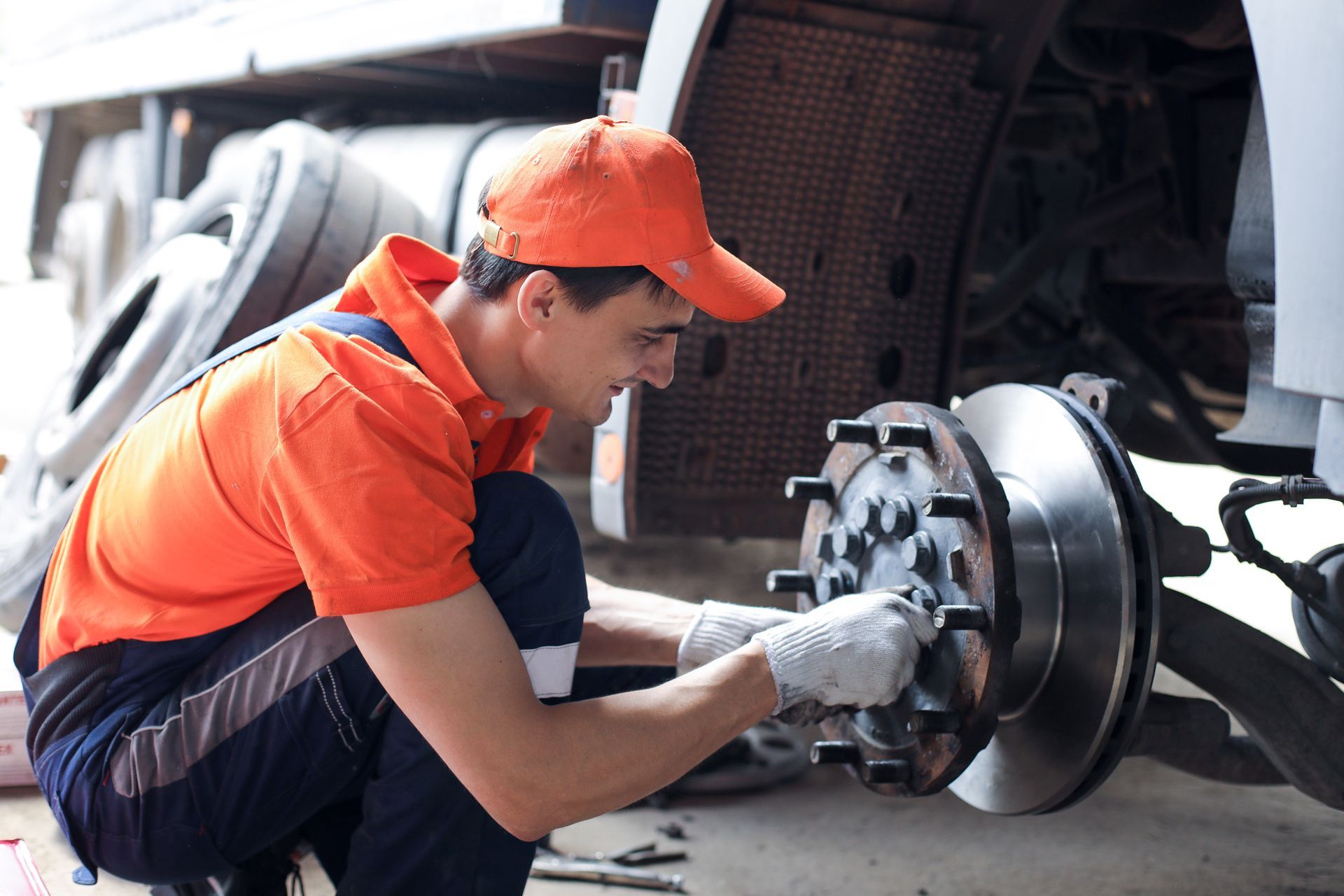 A mechanic in an orange uniform and cap kneels to perform maintenance on a truck wheel assembly.
