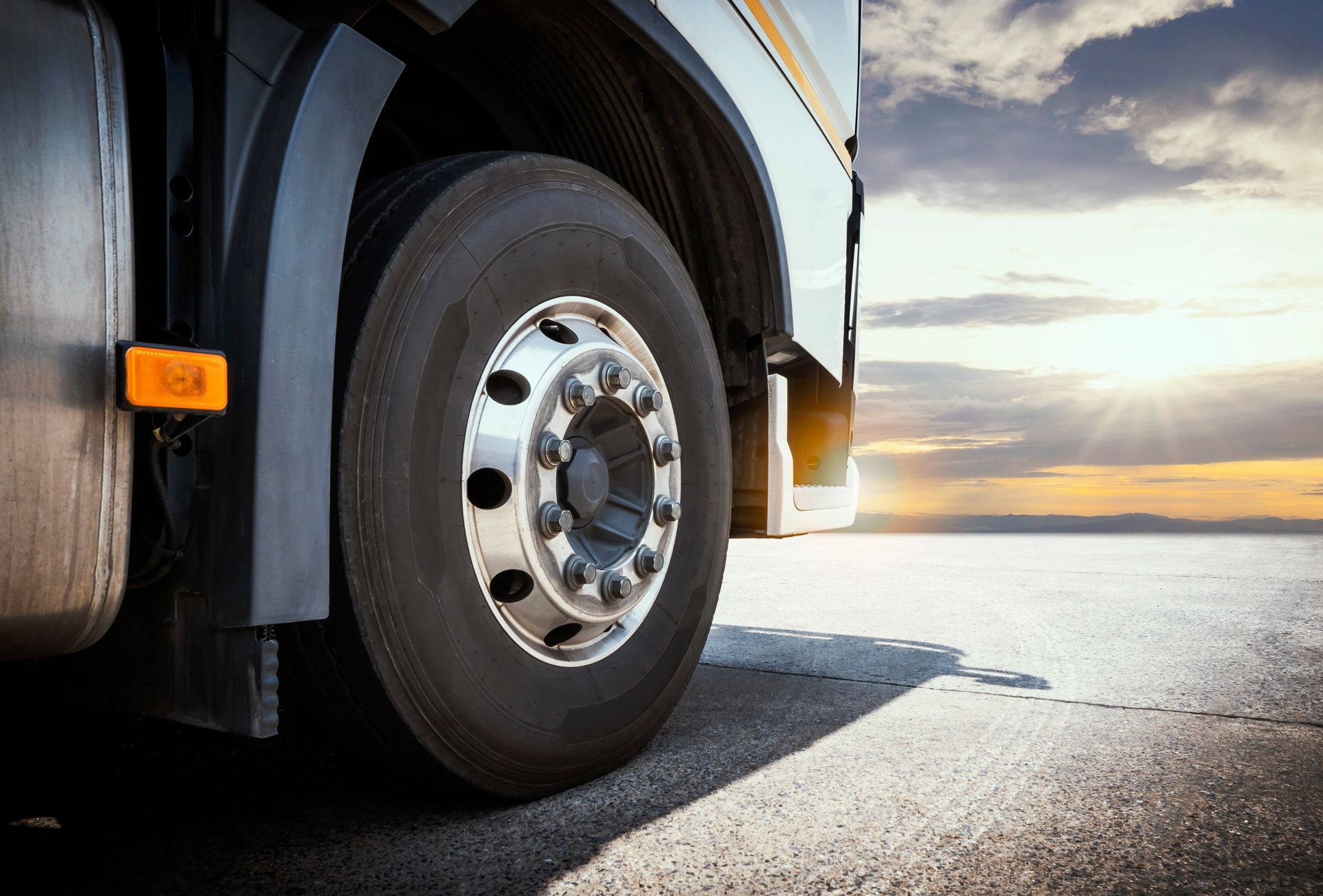A close-up of a large truck tire and wheel on a road against a sunrise sky.