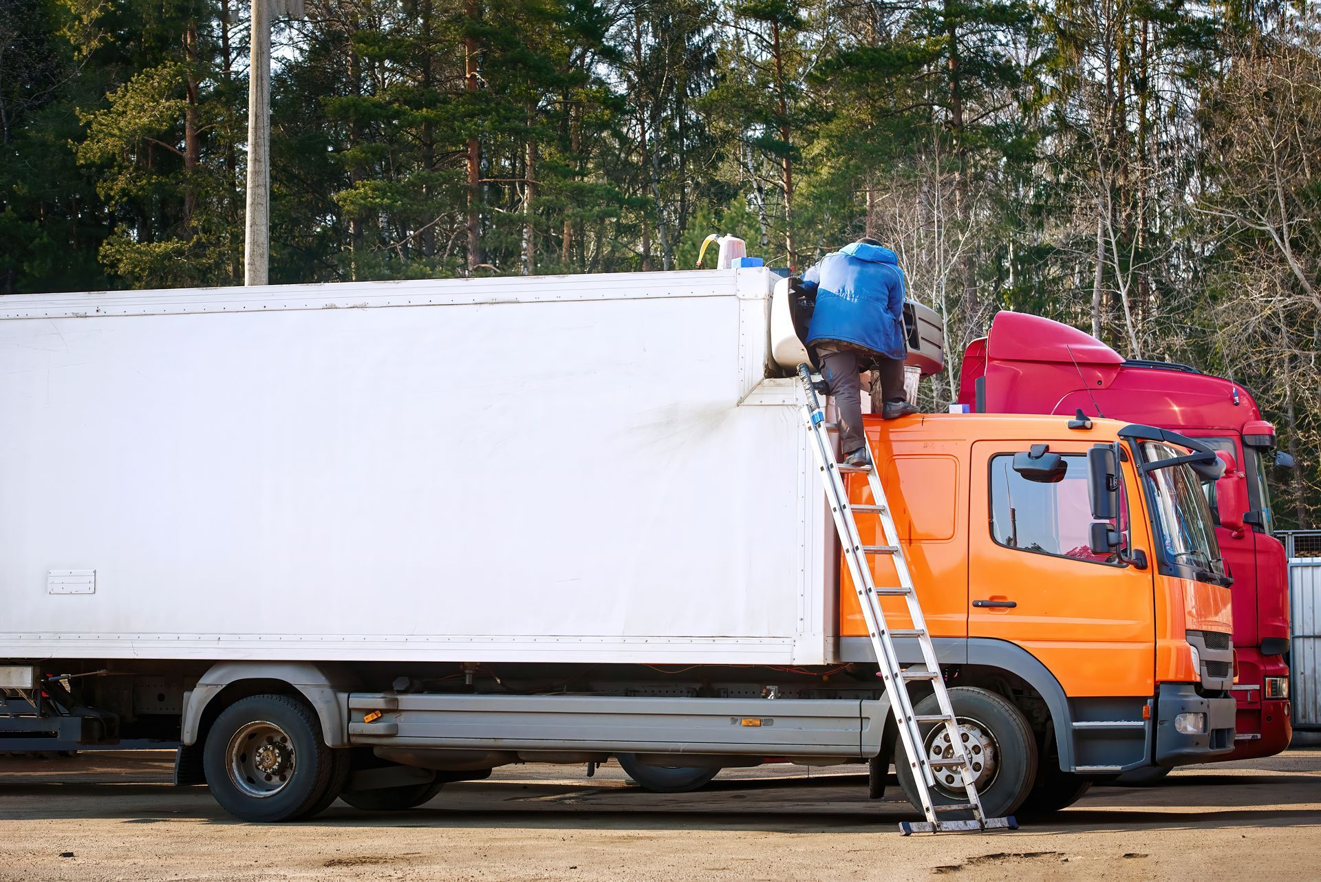 A person in a blue jacket climbs a ladder leaning against the orange cab of a white box truck outdoors.