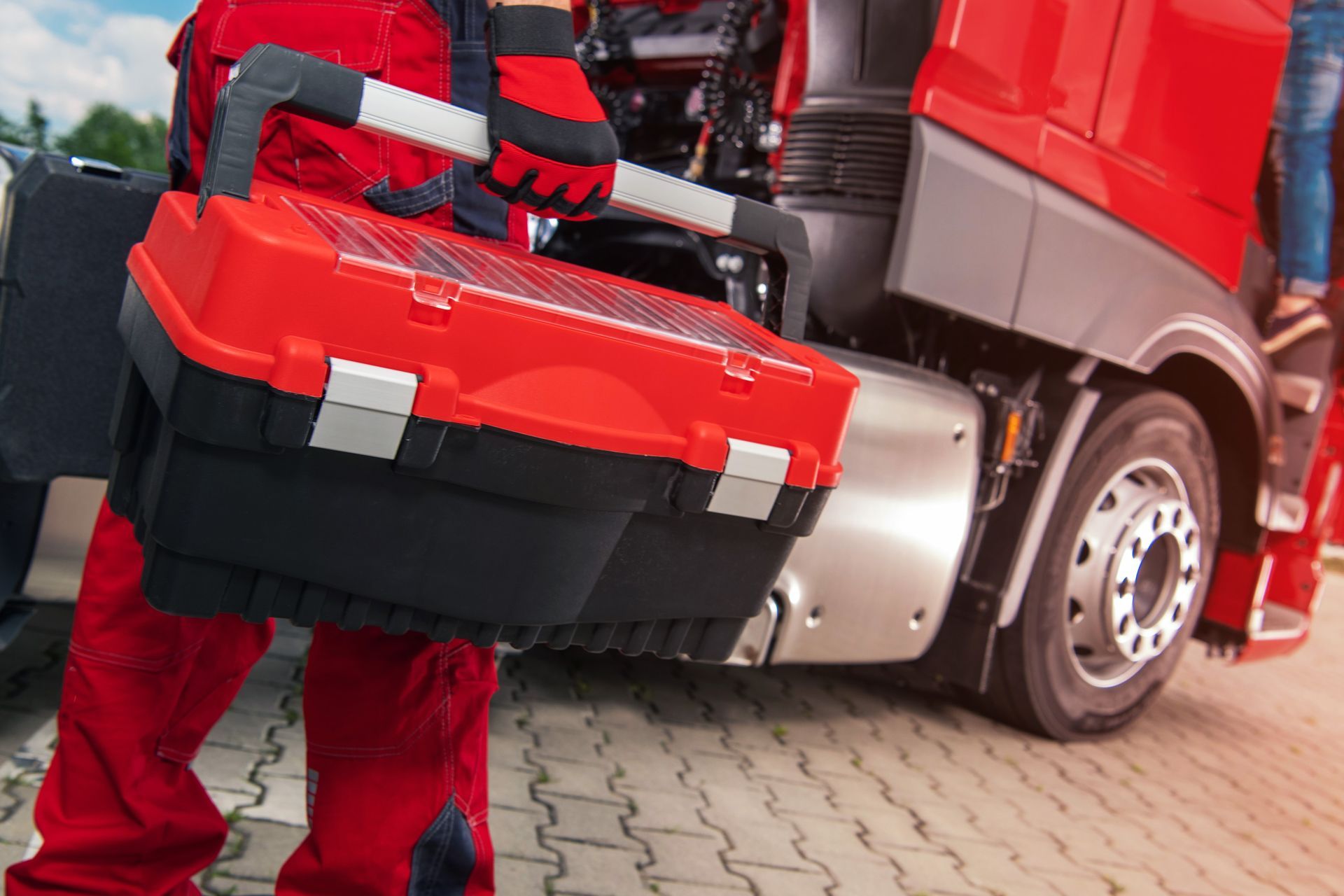 A mechanic in red workwear holds a large red and black toolbox beside a red semi-truck.