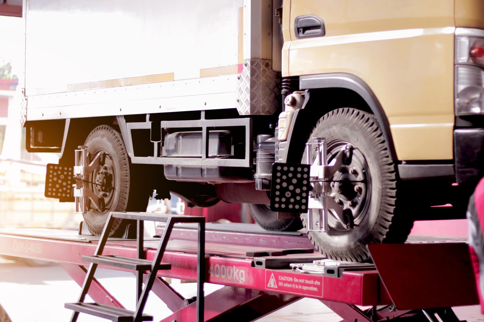 A light-colored delivery truck undergoes a wheel alignment check on a red automotive lift in a workshop.