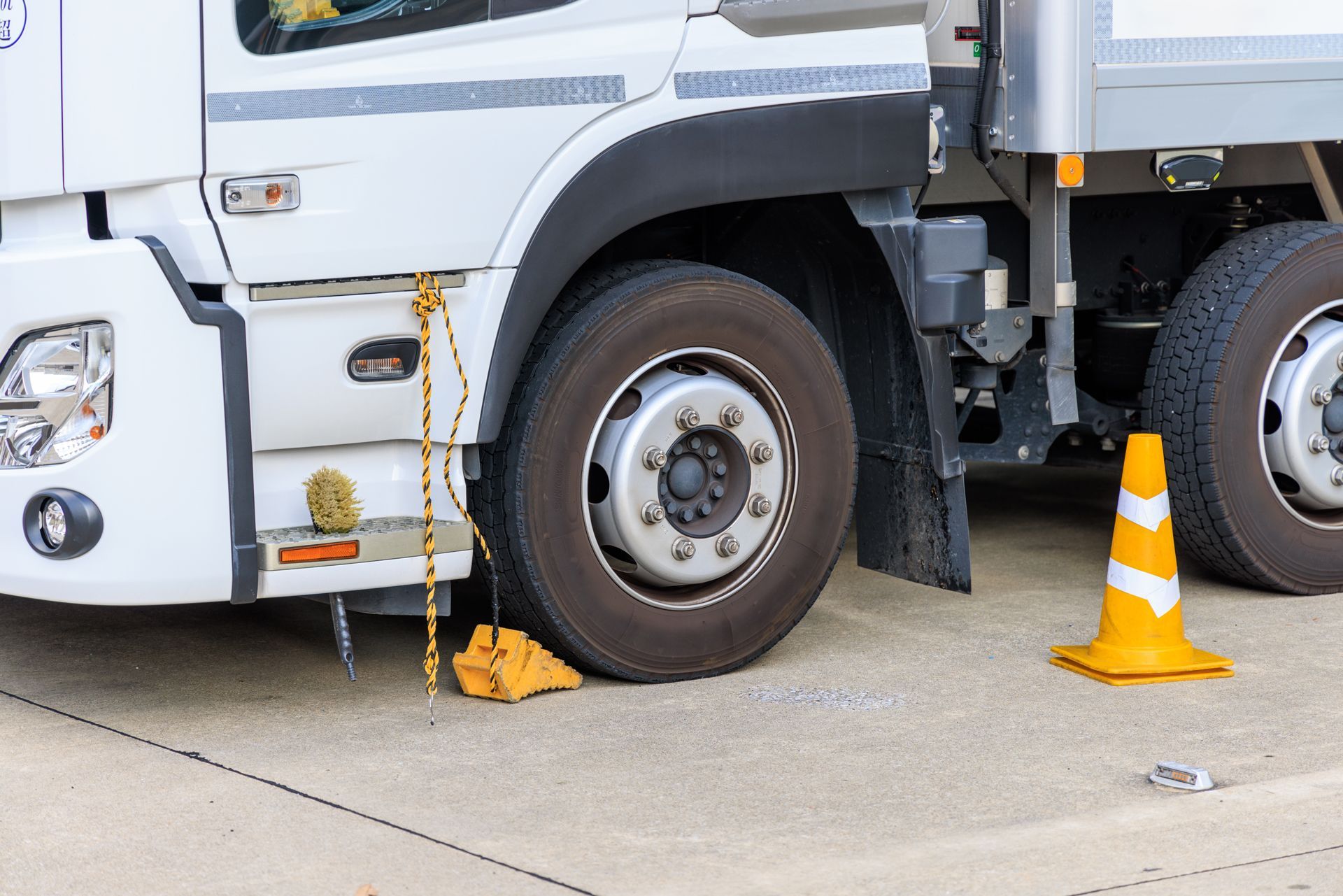 A white truck parked on concrete with a yellow wheel chock and an orange traffic cone placed near the front tire.