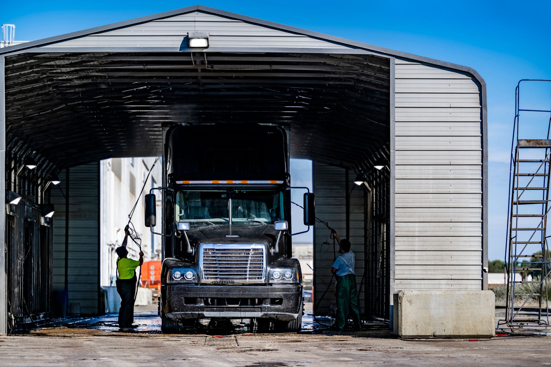 Two workers pressure-wash a black semi-truck parked inside an open-air metal bay under a clear blue sky.