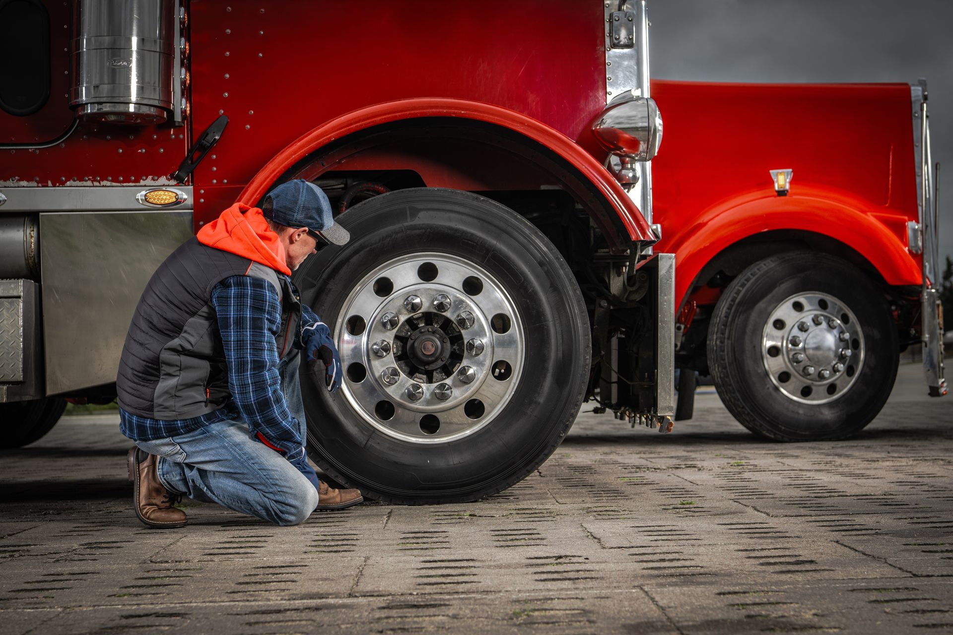 A person kneeling and inspecting the tire of a large red semi-truck parked on a concrete surface.