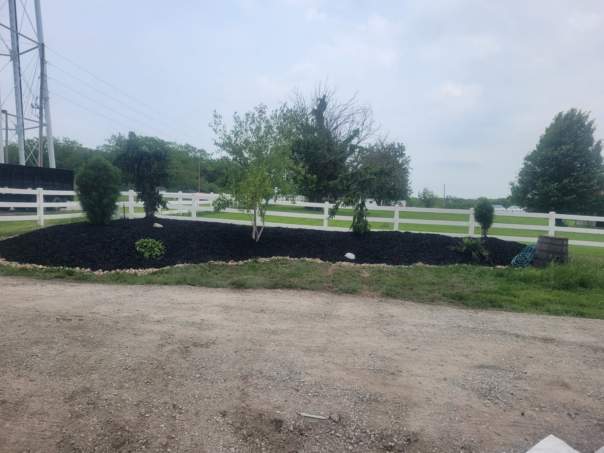 Landscaped garden bed with young trees and shrubs beside a gravel road, white fence in the background