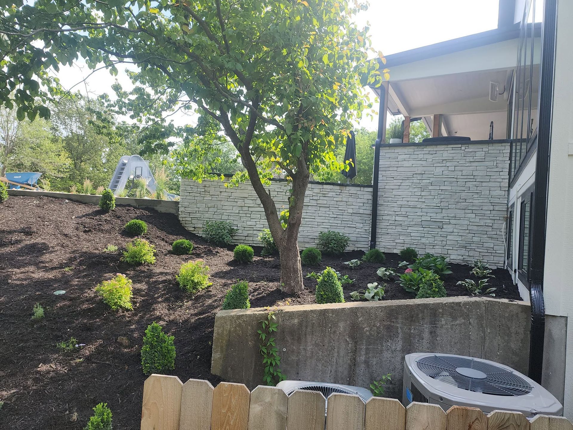 Terraced garden with young plants, a tree, stone retaining walls, and a house patio with an air conditioner unit