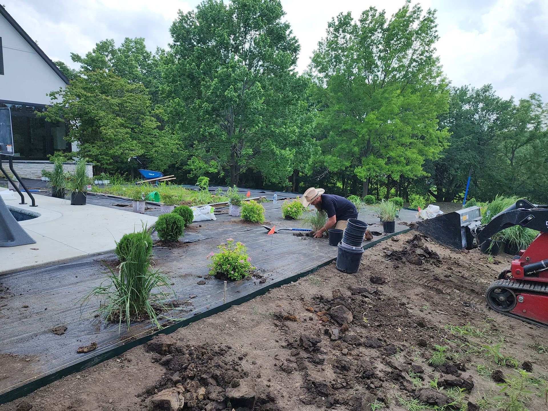 Backyard landscaping with workers, mulch, shrubs, and a red wheelbarrow by a house