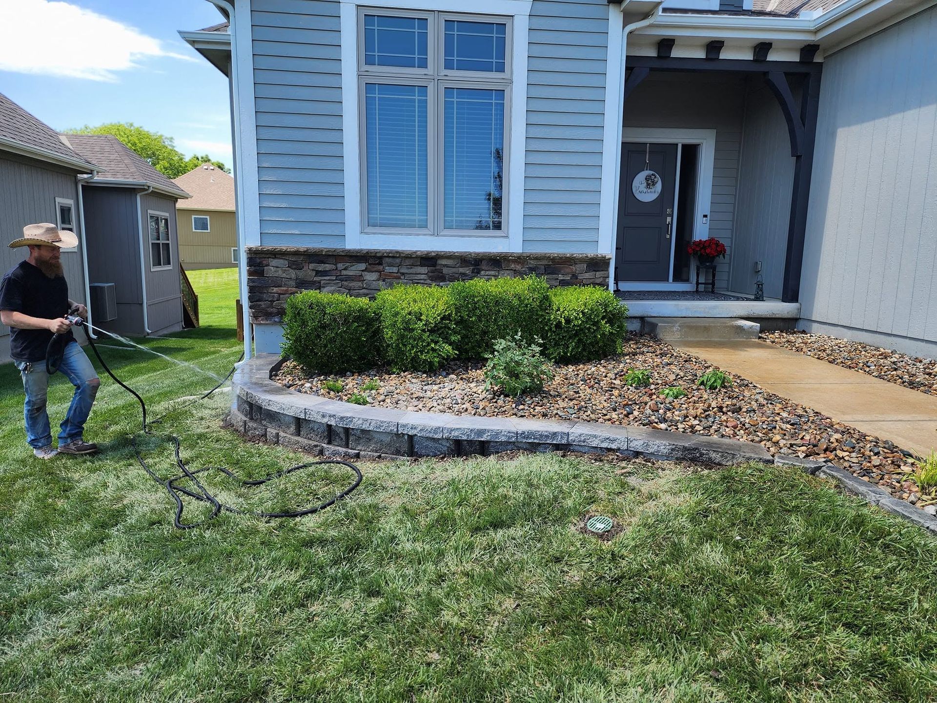 Person spraying a house foundation and lawn with a hose near a front porch and window.