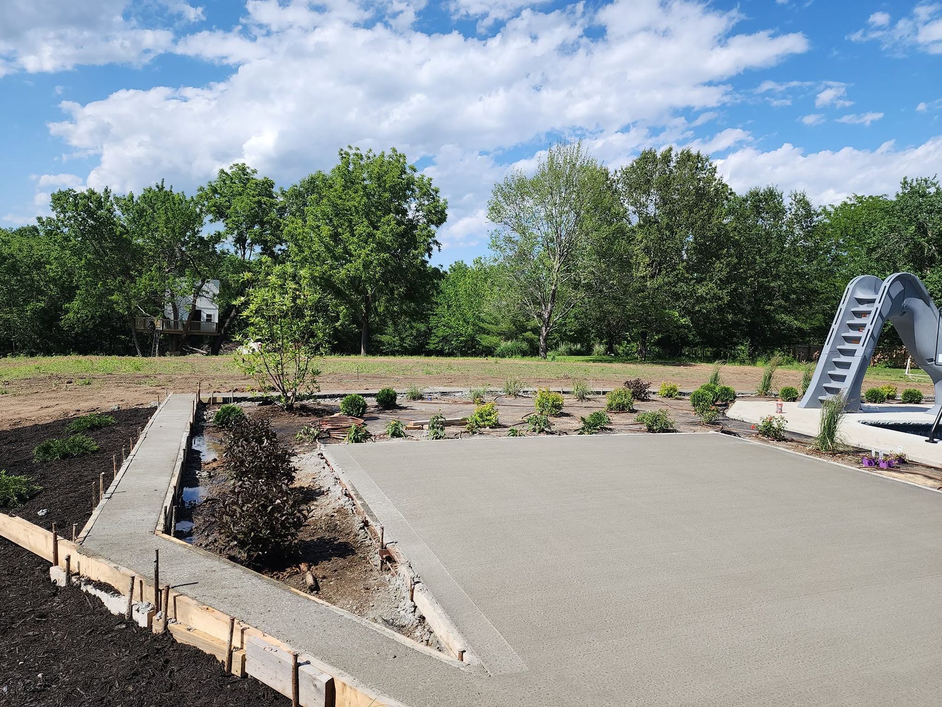 Outdoor garden area with a concrete patio, young plants, trees, and a blue playground slide under a partly cloudy sky