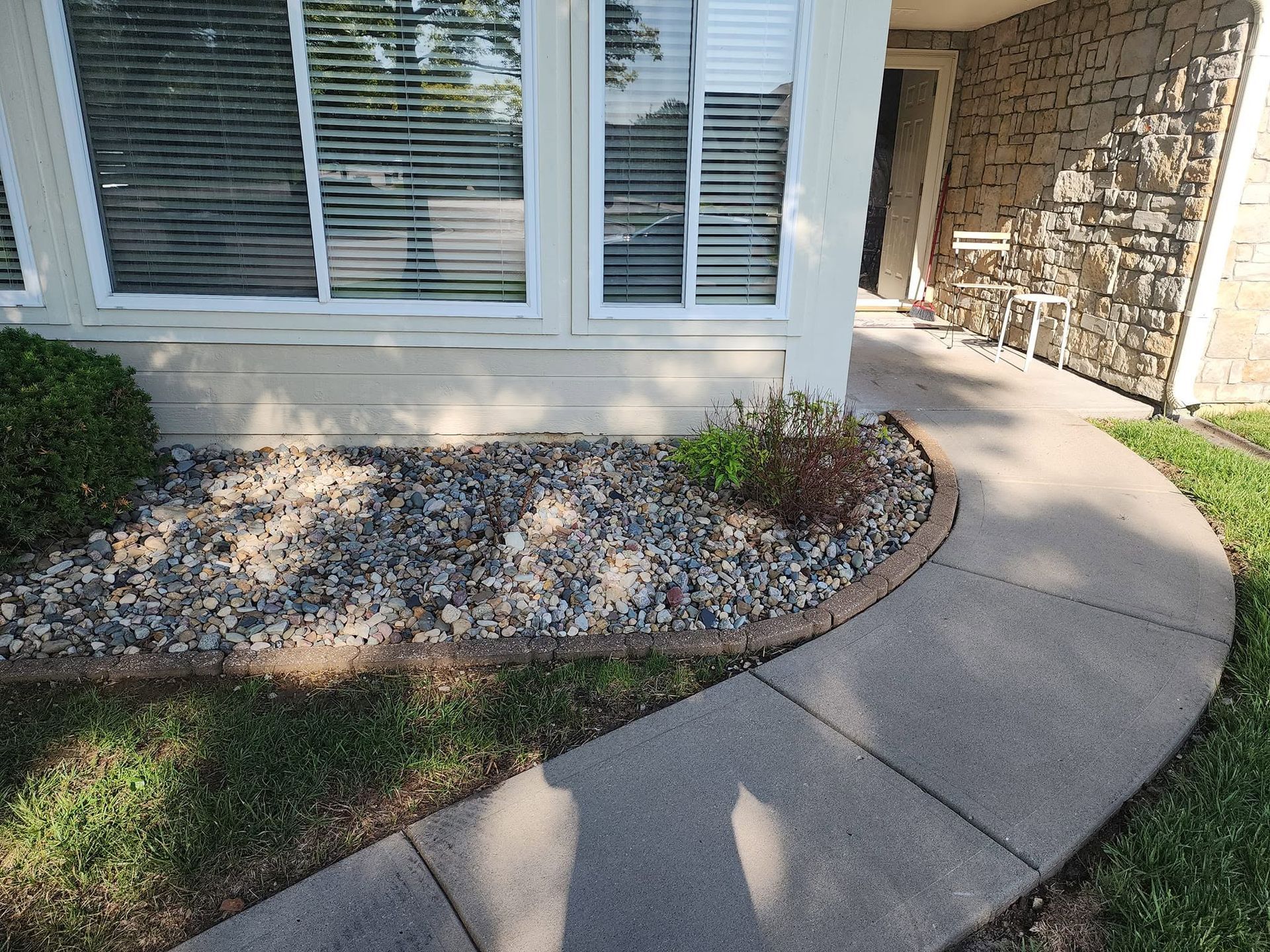 Curved concrete walkway beside a house window and stone entry, bordered by gravel and a small shrub bed