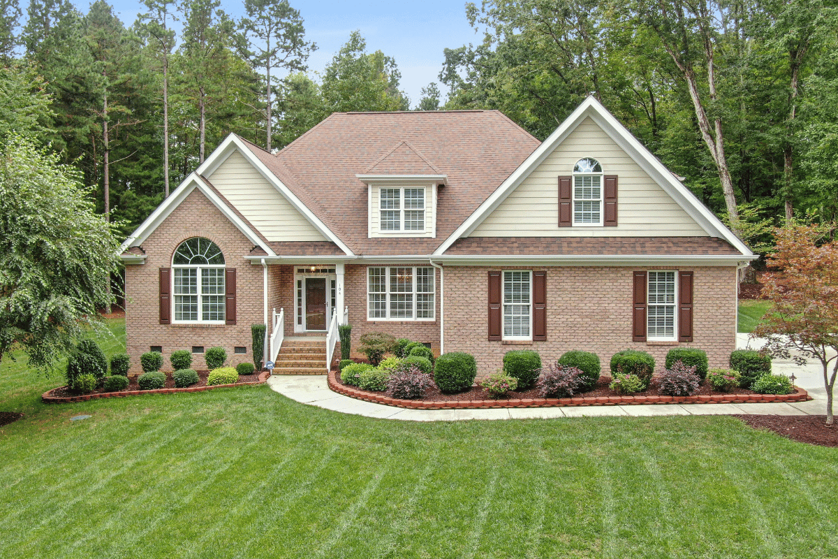 Brick suburban house with beige siding, red shutters, and a manicured green lawn, fronted by trees