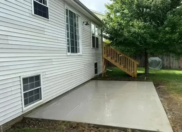 Gray concrete patio next to a house with white siding and a wooden staircase leading up to a deck.