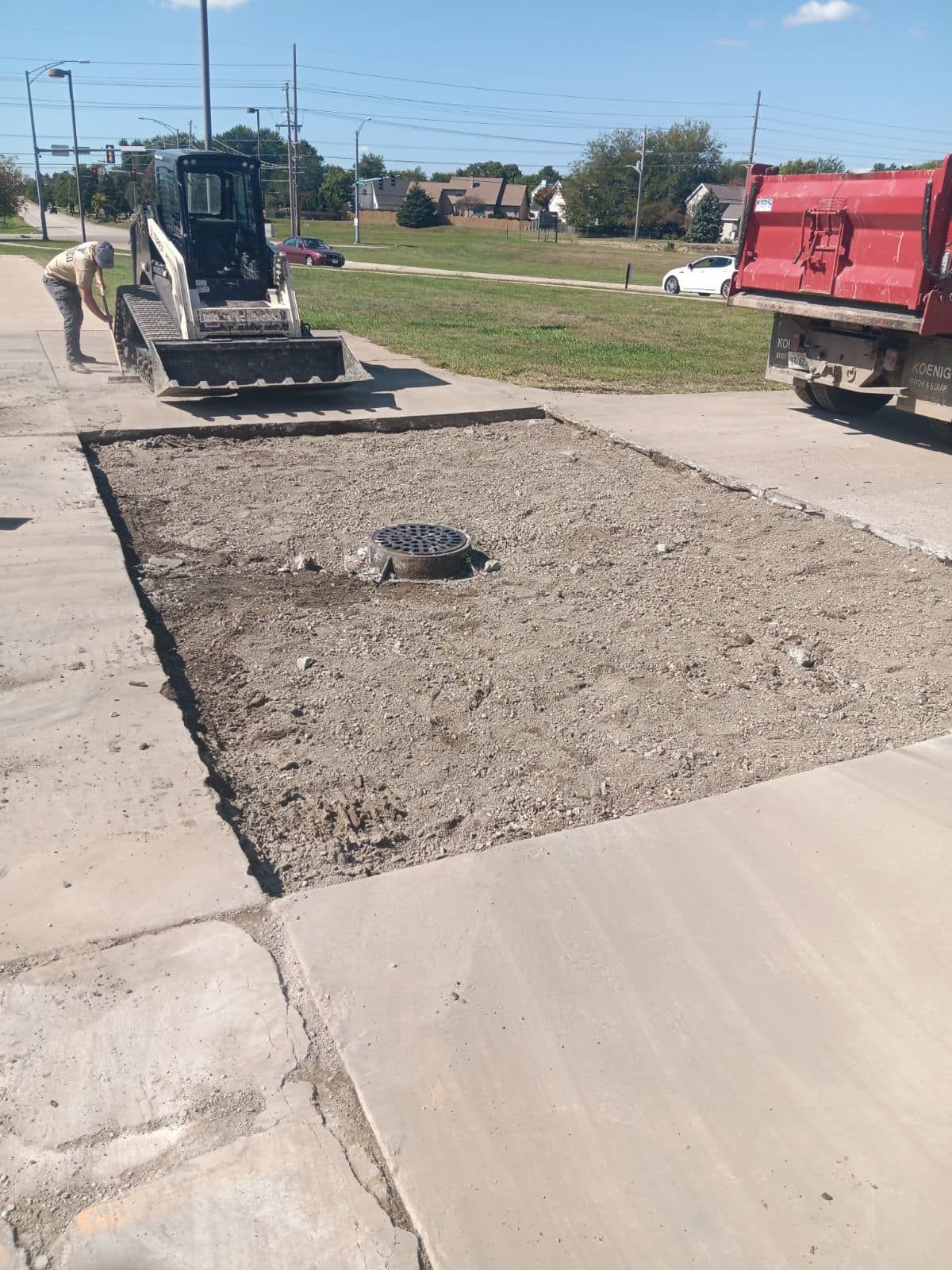 Sidewalk repair: a small skid steer and worker next to a section of removed concrete. Red truck in background.