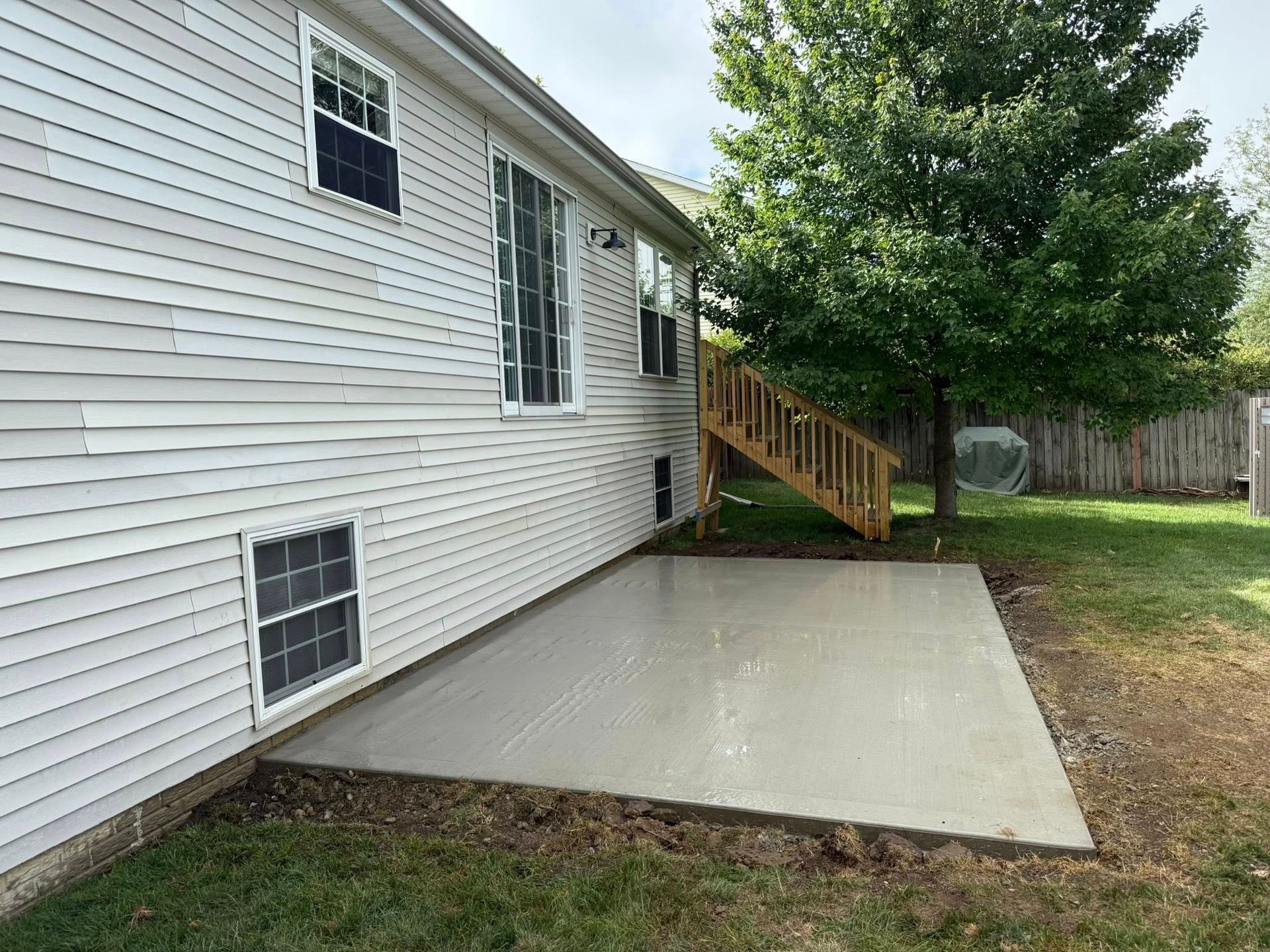 Gray concrete patio next to a house with white siding and a wooden staircase leading up to a deck.