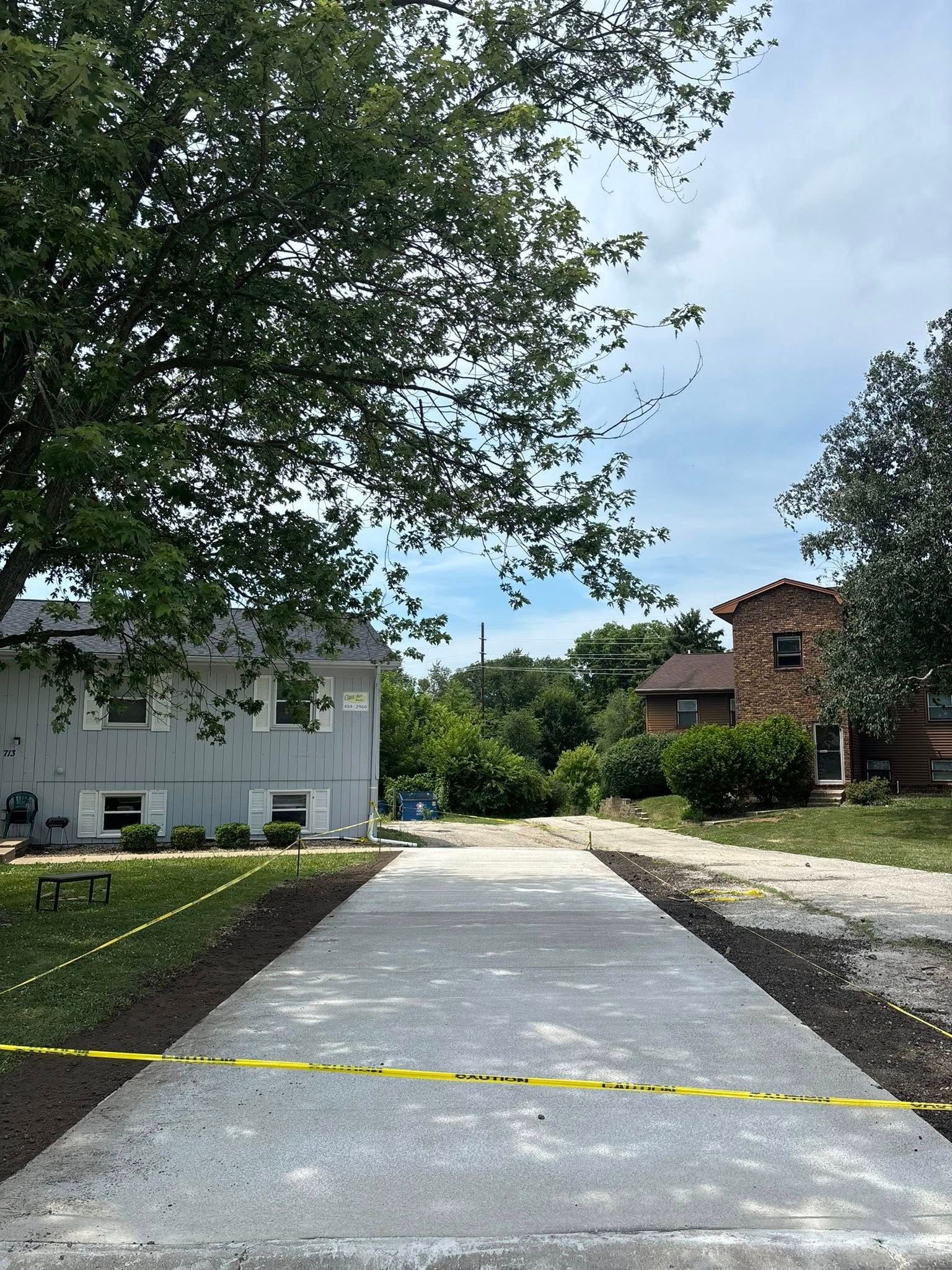 Newly poured concrete driveway with caution tape, houses, and trees. Overcast sky.