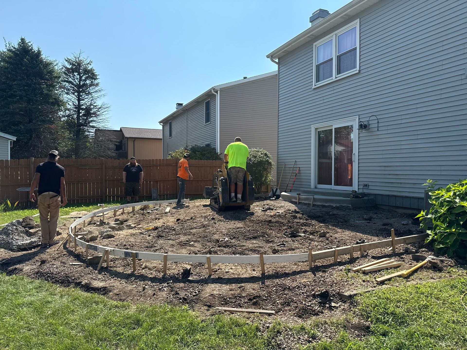 Construction site in backyard. Workers shaping dirt with equipment, near a house and a wooden fence.