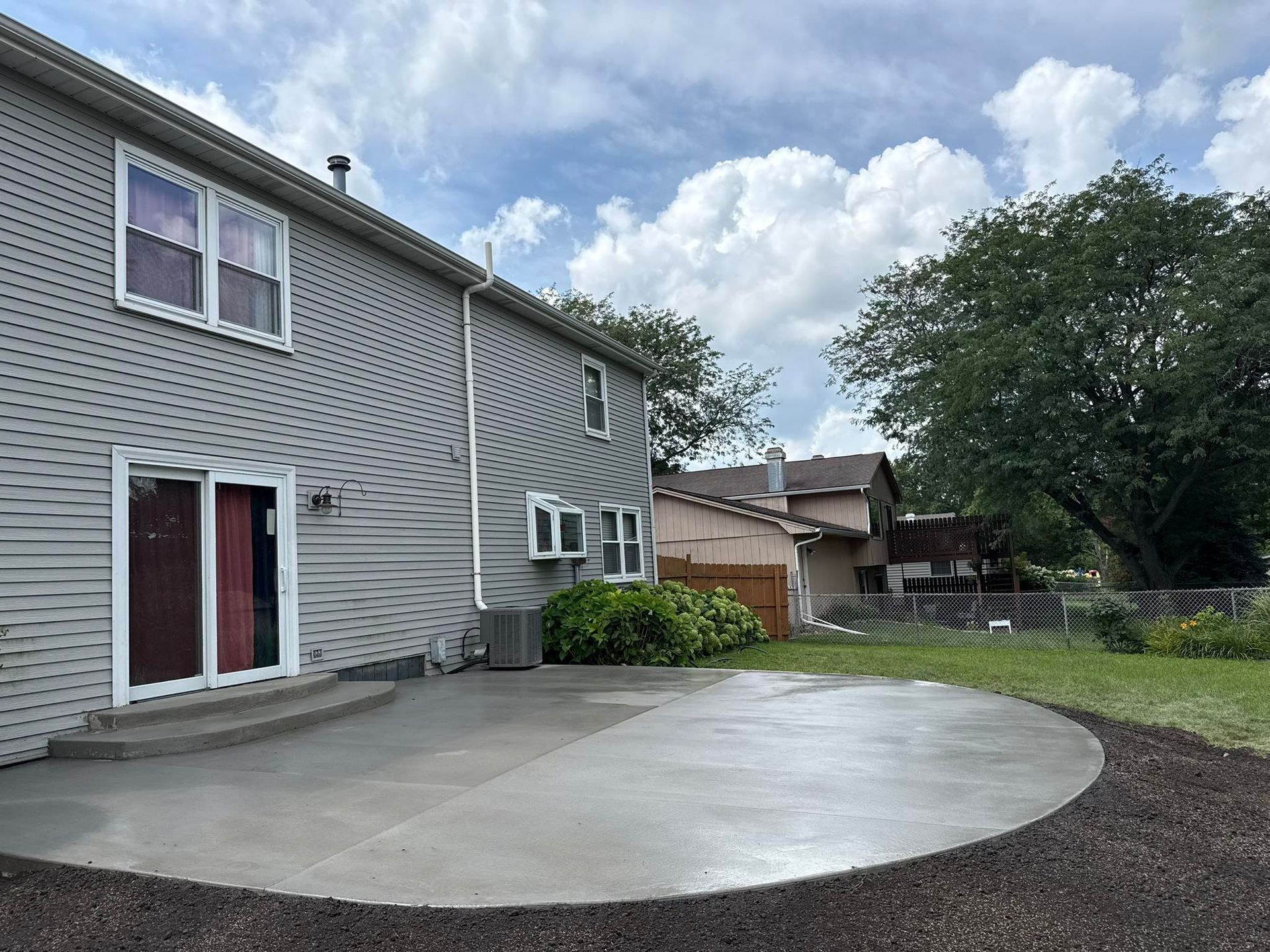 Backyard with a concrete patio beside a two-story gray house. A sunny day with a cloudy sky.