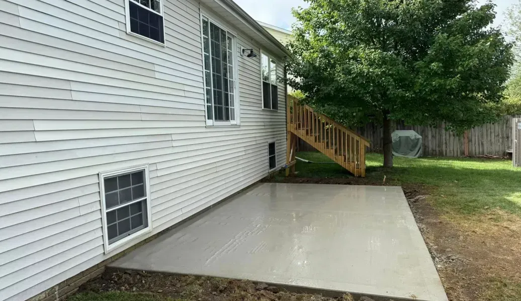 Side view of a house with a concrete patio. Stairs lead to a wooden deck. A tree and grass surround the house.