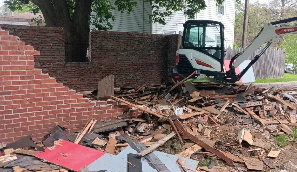 Bobcat machine demolishing brick wall by tree, surrounded by debris.