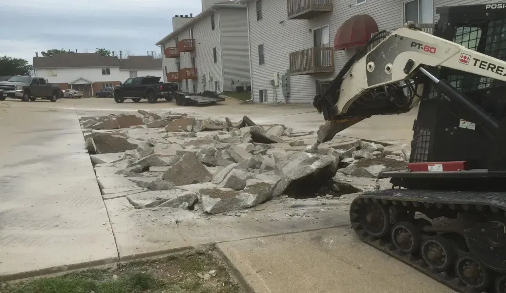 Construction vehicle demolishing concrete in front of an apartment building on a cloudy day.