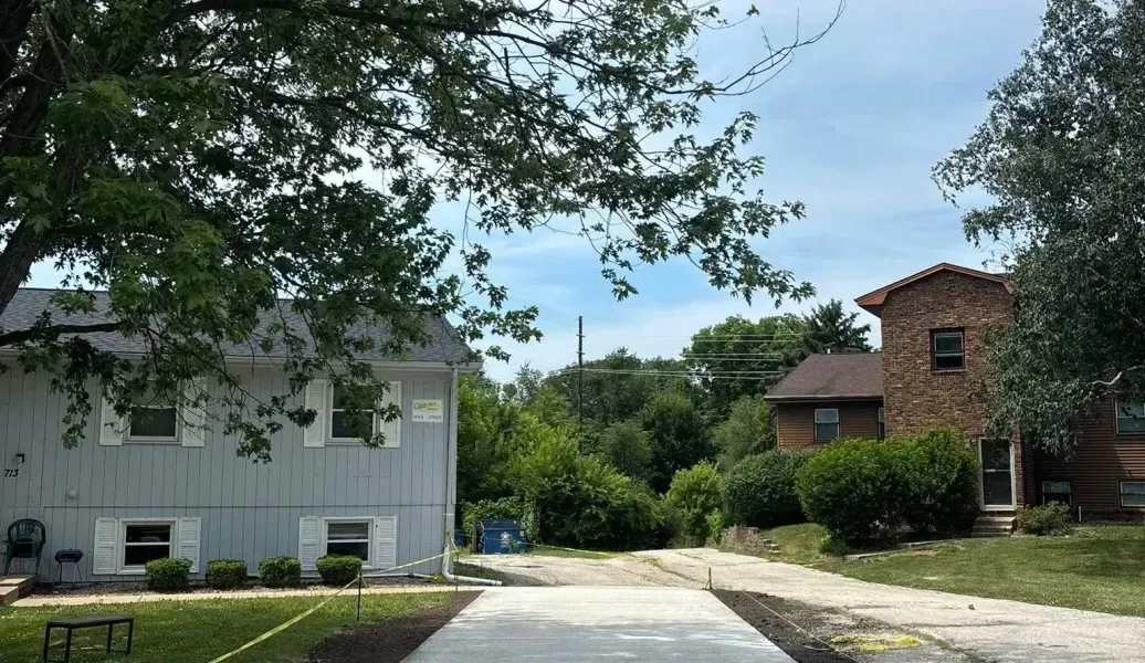 Newly paved concrete driveway leading towards houses under a cloudy sky, with caution tape and trees.