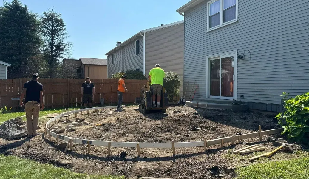 Construction workers building a curved concrete border in a backyard. A small excavator is in use.