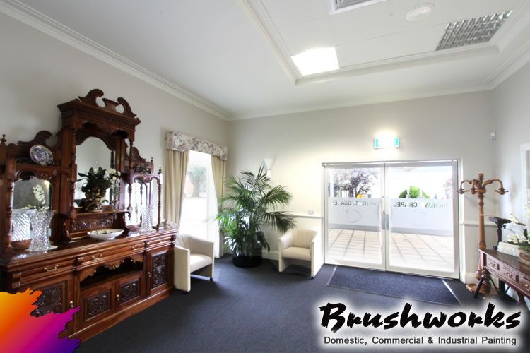 A Man is Standing on a Ladder Painting a Wall With a Paint Roller — Brushworks Painting In North Albury, NSW.