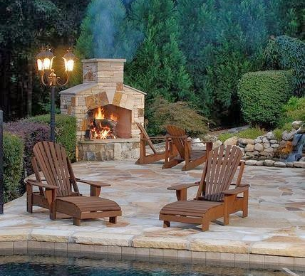 Outdoor fireplace with Adirondack chairs on a stone patio near a pool, surrounded by greenery. Outdoor fireplace with Adirondack chairs on a stone patio near a pool, surrounded by greenery.