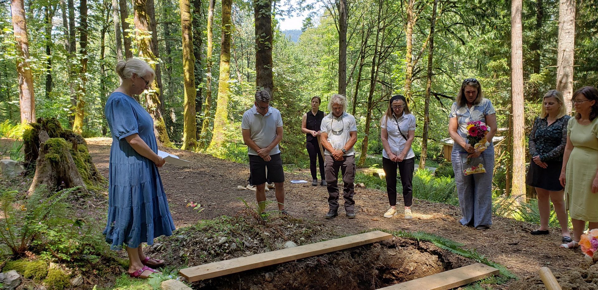 Scene at a Natural Burial site in a West Coast Forest setting. Eight people are standing around the open grave. Boards are edging the grave.  Salt Spring Island Natural Cemetery.  