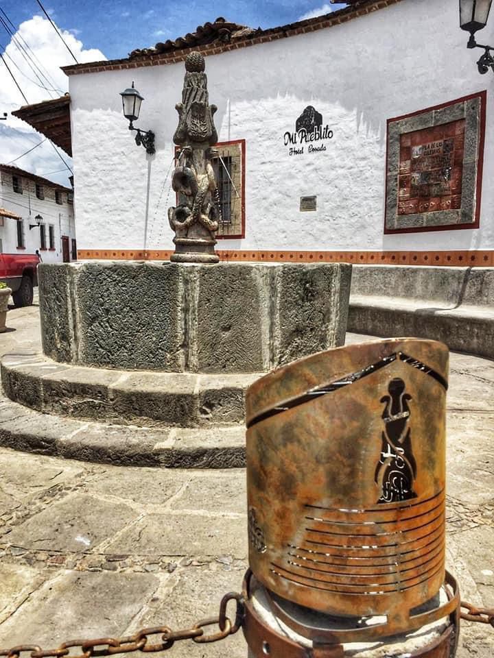 Una fuente de piedra con un pilar tallado en una plaza empedrada frente a un edificio blanco con un mural ornamentado.