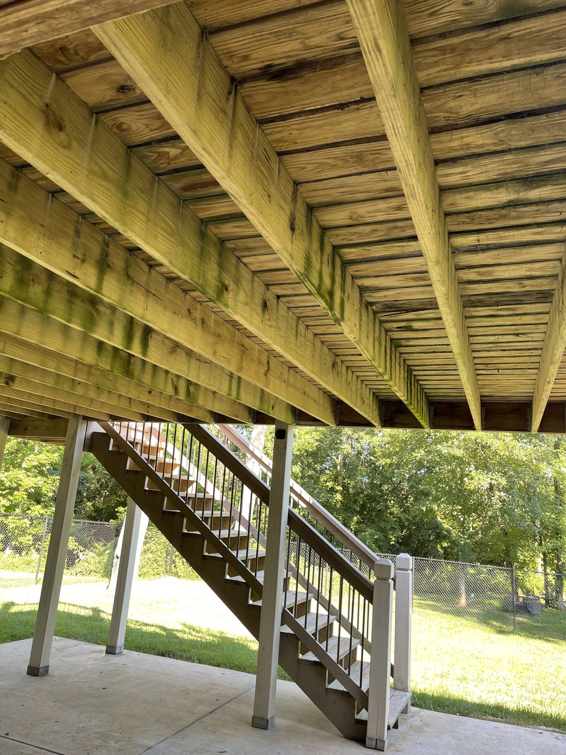 Under-deck view of a wooden staircase leading to a raised deck, with trees and grass beyond
