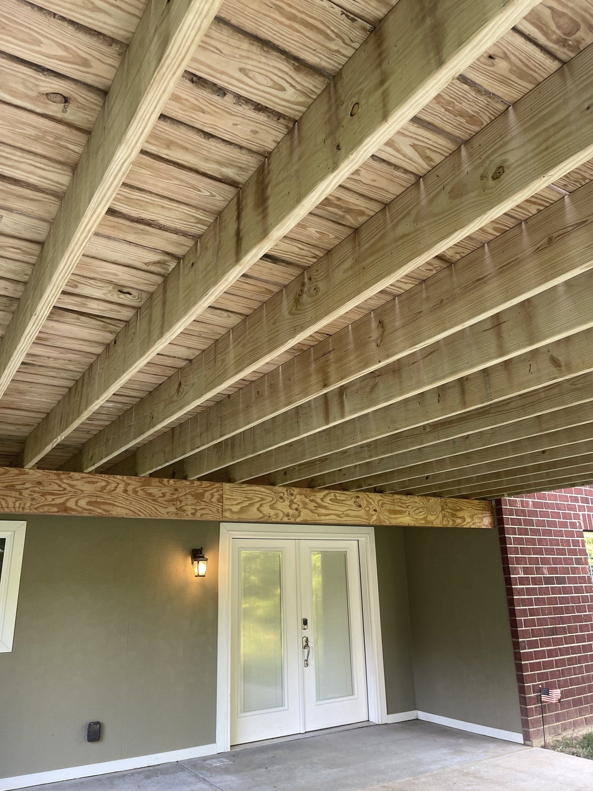 Covered patio with exposed wood beams, gray wall, white French doors, and brick column under a deck