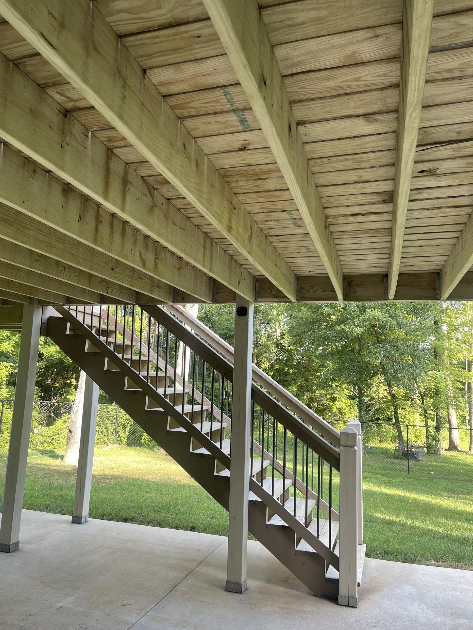 Stairs under a raised wooden deck beside a grassy yard with trees and sunlight