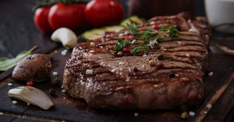 A large steak is sitting on a wooden cutting board surrounded by vegetables.