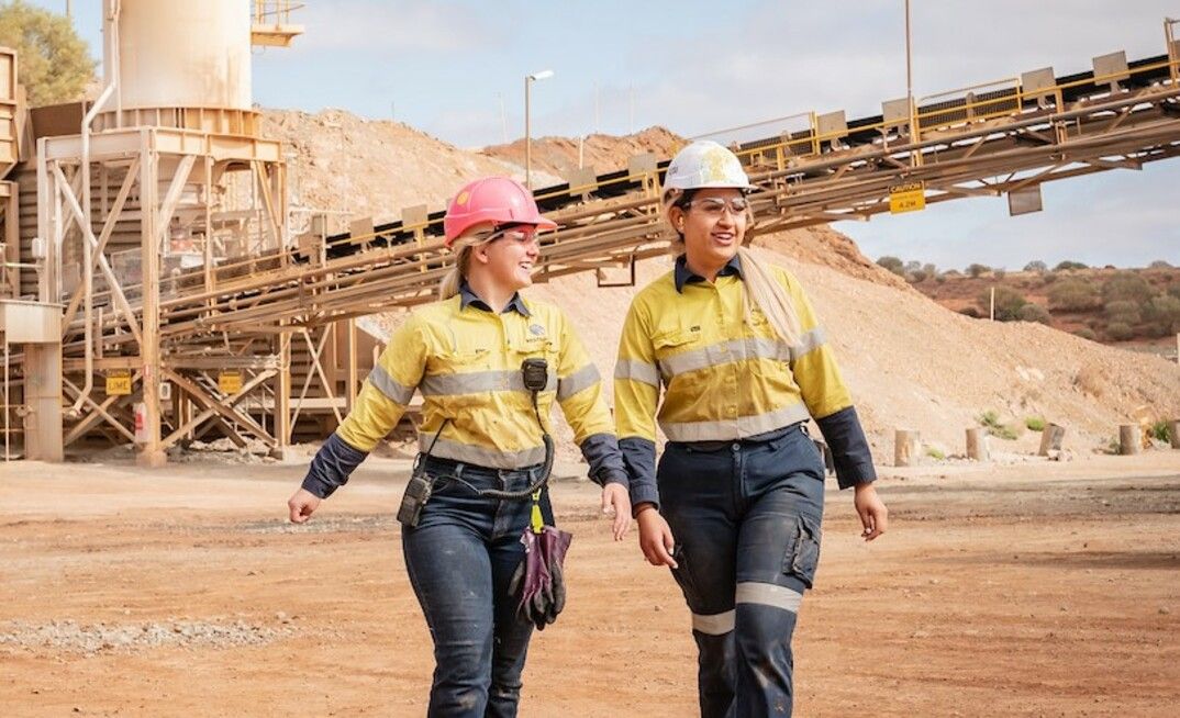 Two women are walking in a dirt field in front of a conveyor belt.