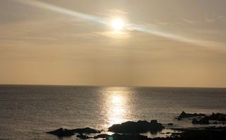 The sun is setting over the ocean with rocks in the foreground.