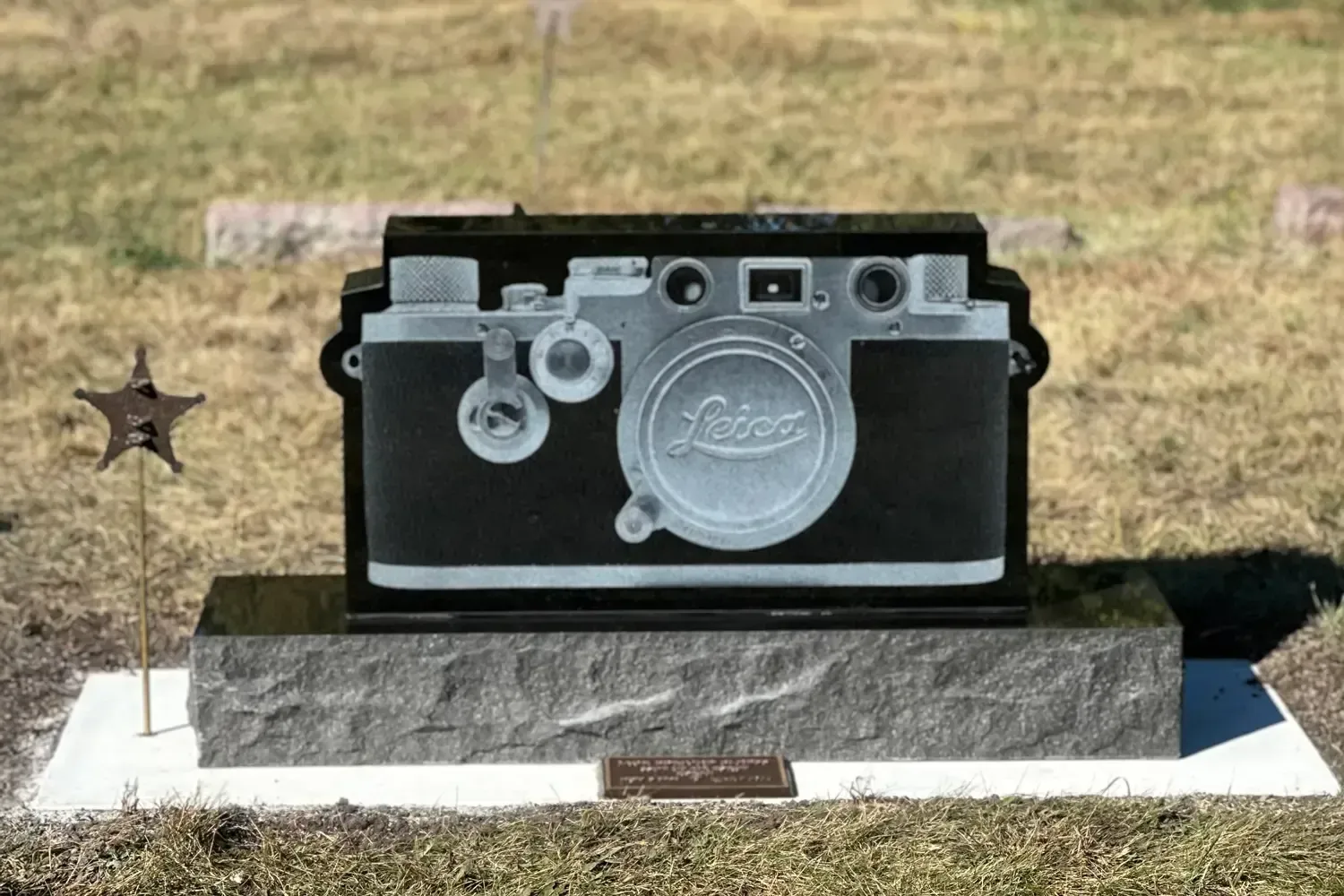 Stone memorial with a large camera-shaped monument on a grassy grave marker