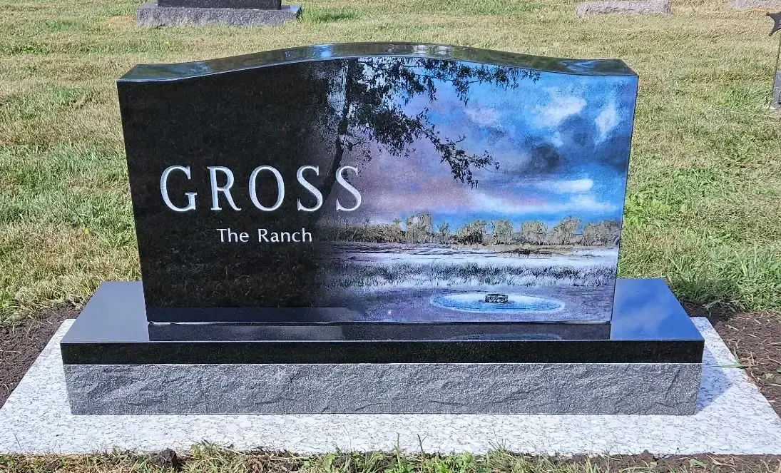 Black granite memorial headstone with “CROSS” and a scenic lake engraving in a grassy cemetery.