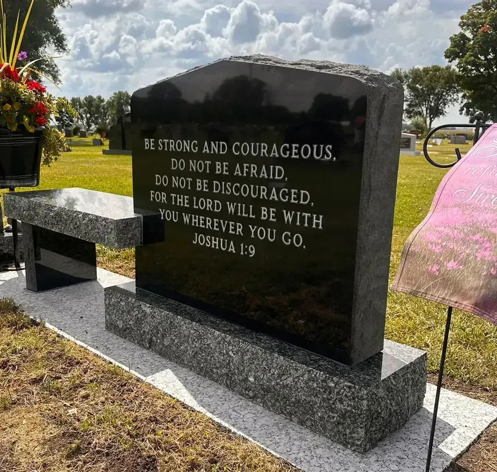 Black memorial stone with Bible verse in a grassy cemetery, flowers beside it.