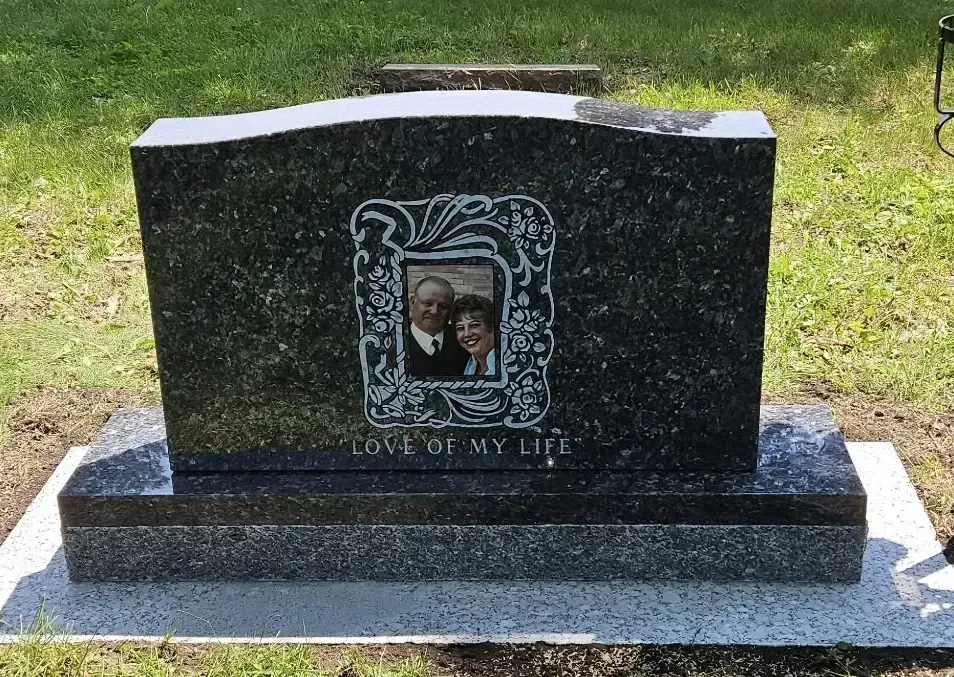 Black granite gravestone with a small inset photo, set on a concrete base in a grassy cemetery.