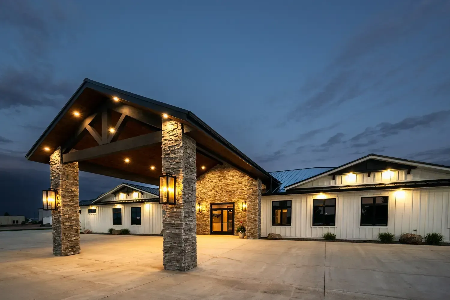 Lit stone building entrance at dusk with warm lights and a large covered driveway
