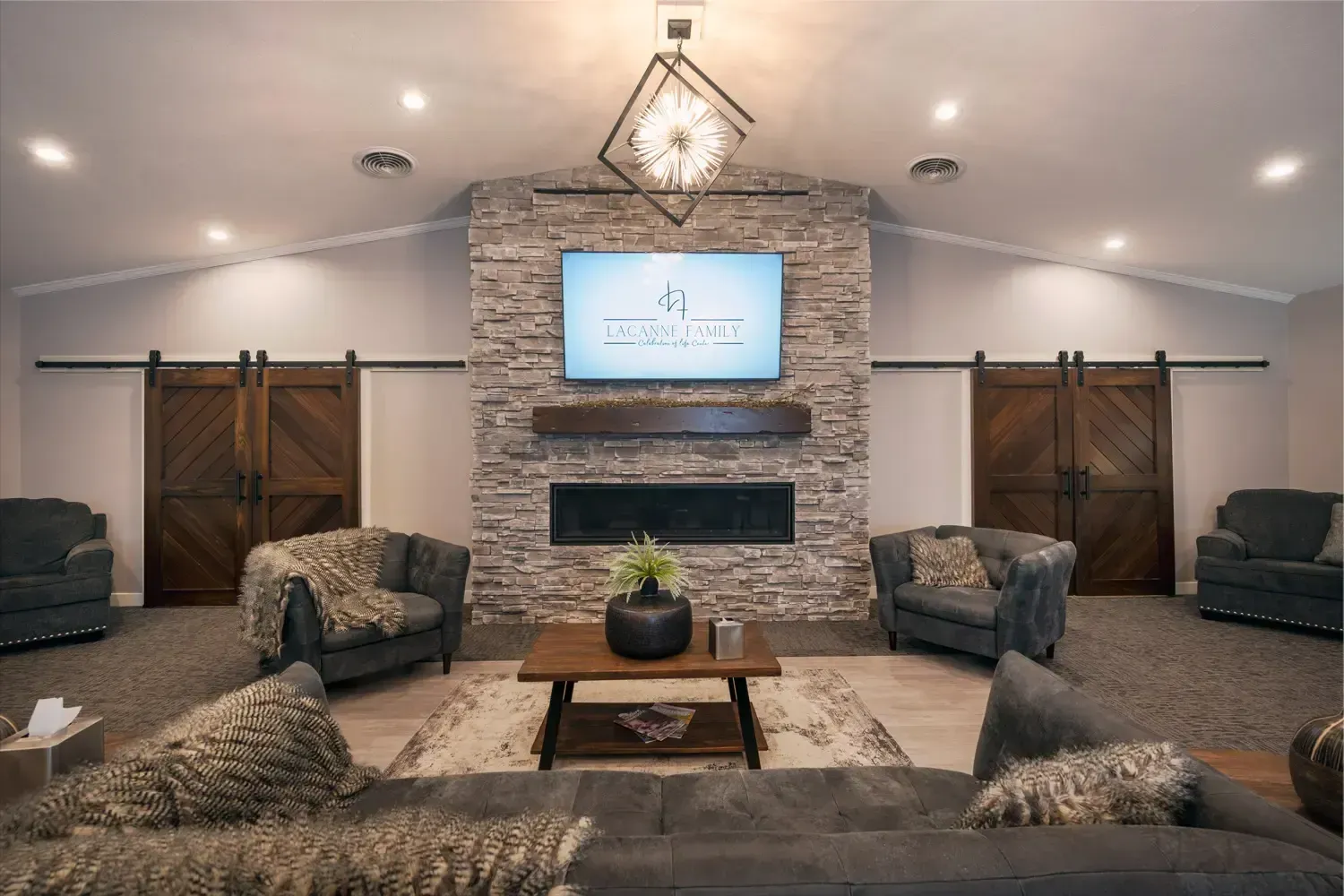 Cozy living room with stone fireplace, TV, wooden coffee table, and gray sofas under warm recessed lighting