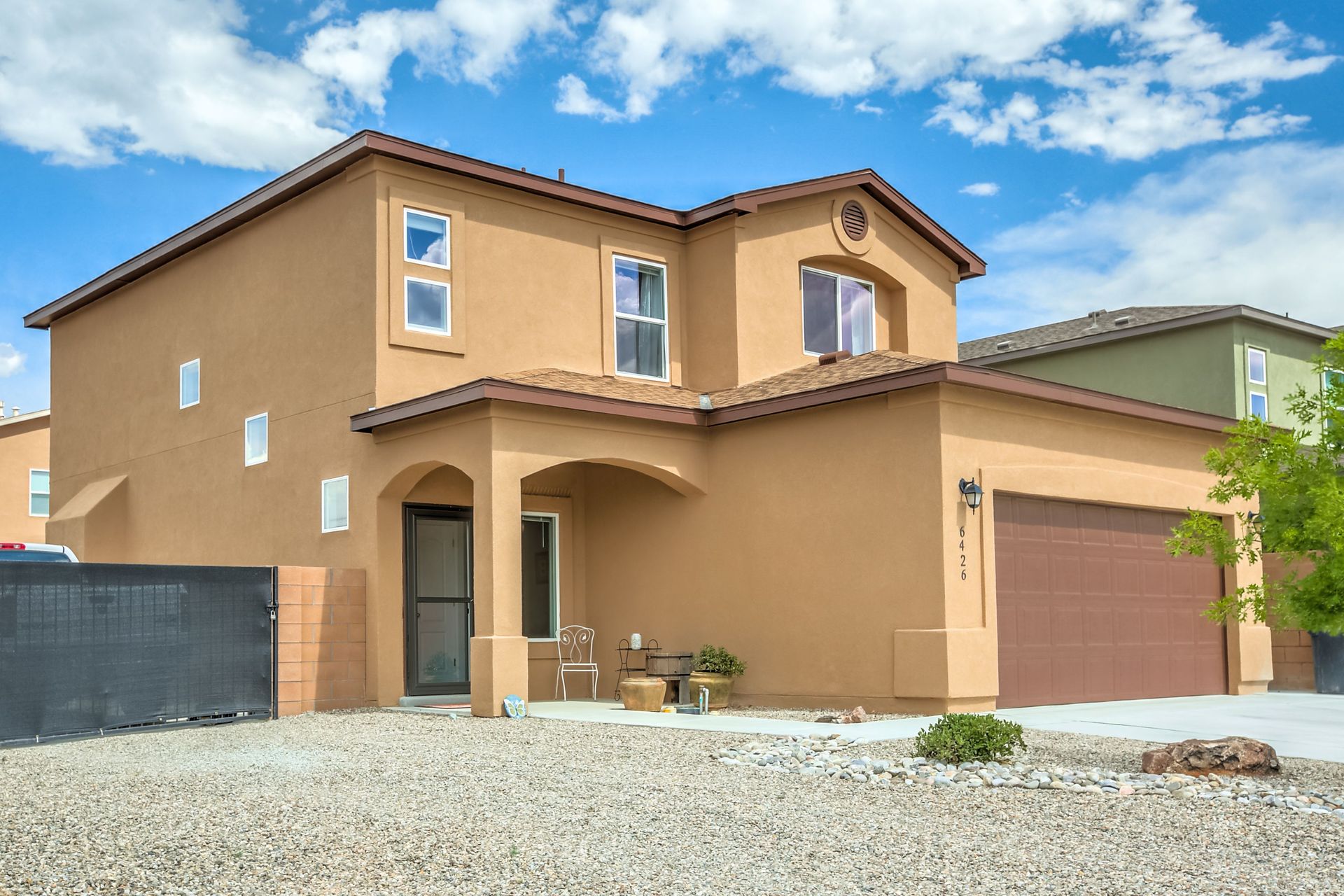 Tan stucco two-story house with a red garage door and gravel front yard under a blue sky