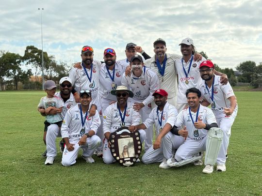 Cricket team in white uniforms posing on a grassy field with medals and a trophy