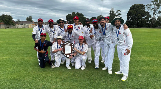 Group of cricketers in white uniforms and red caps posing on a grassy field, holding a trophy.