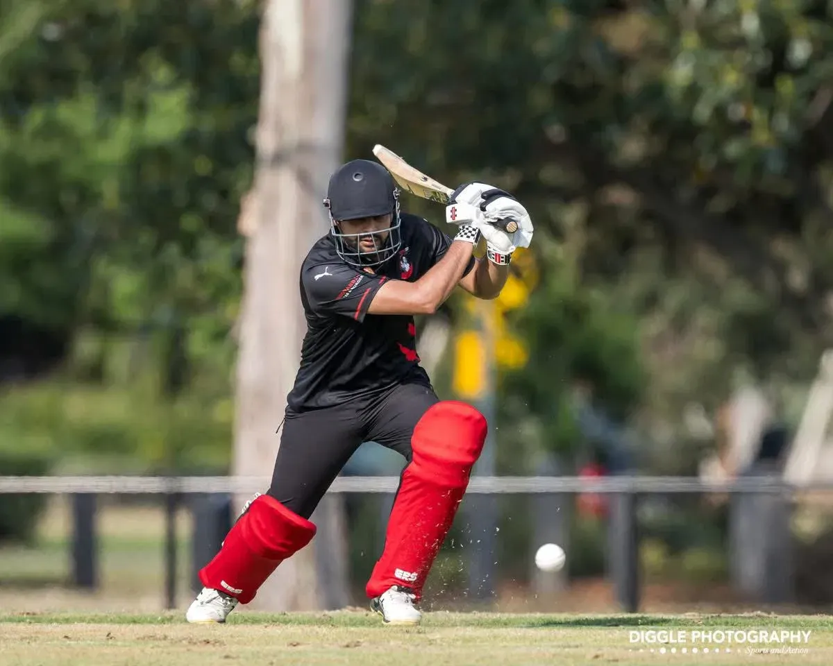 Baseball batter in black and red uniform swings at a pitch on a grassy field.