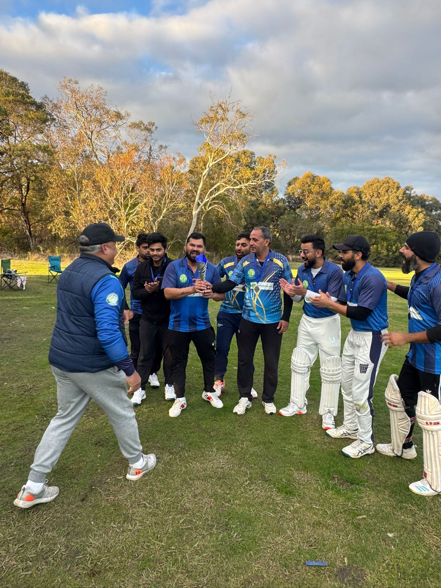 Cricket team in blue jerseys gathers on a grassy field while a coach speaks, with autumn trees behind them.