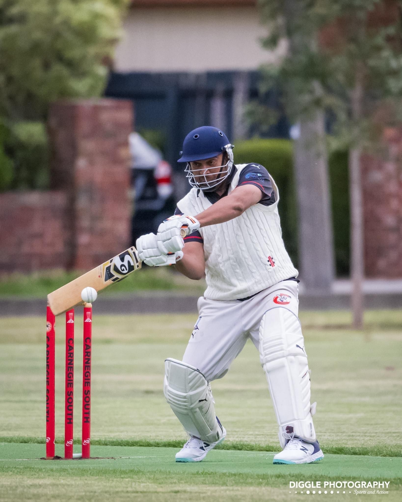 Cricket batter in white swings at the ball near red stumps on a grassy field