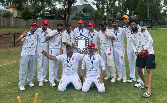 Cricket team in white uniforms posing on a grassy field, some wearing red caps and medals.