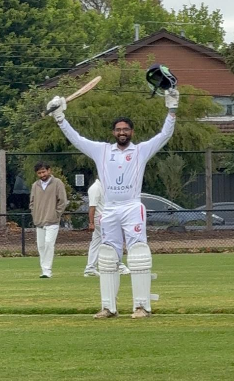 Cricketer in white raises bat and helmet in celebration on a grassy field with a teammate behind him.